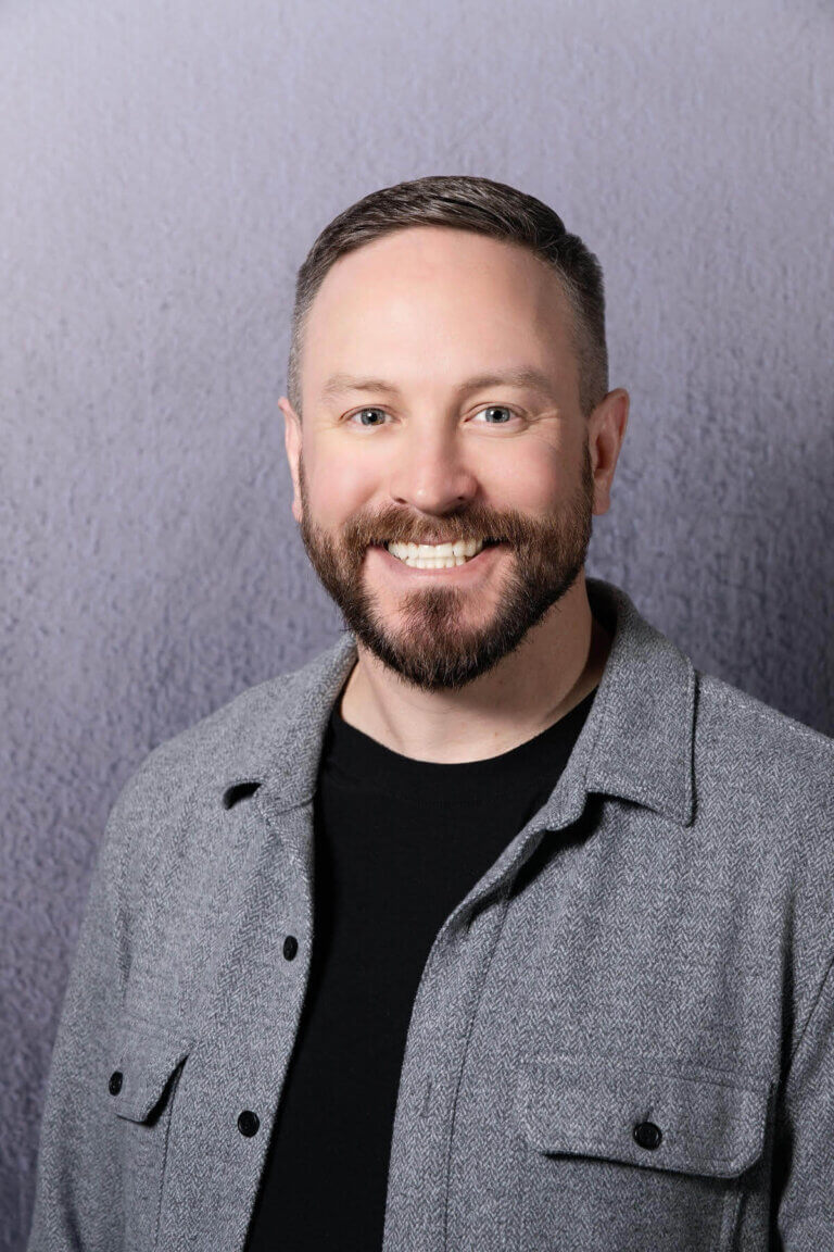 Smiling man with short hair and beard wearing a gray button-up shirt over a black t-shirt, standing in front of a textured gray background.
