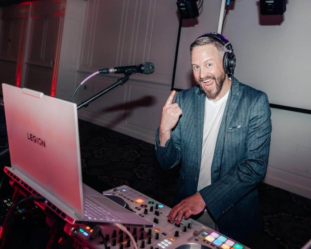 A man wearing headphones stands behind ROQ Events DJ equipment and a laptop, smiling and pointing at himself in a dimly lit room.