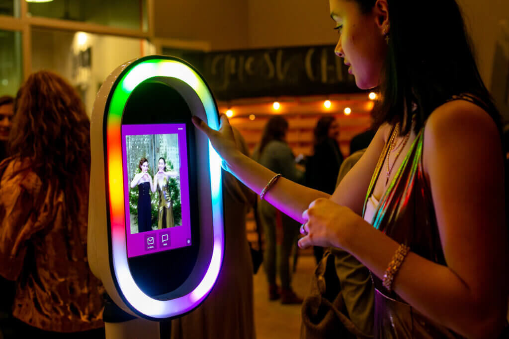 A woman uses a photo booth with a colorful LED frame at an indoor event. The screen displays her photo with another person in front of a decorated backdrop. Other attendees are in the background.