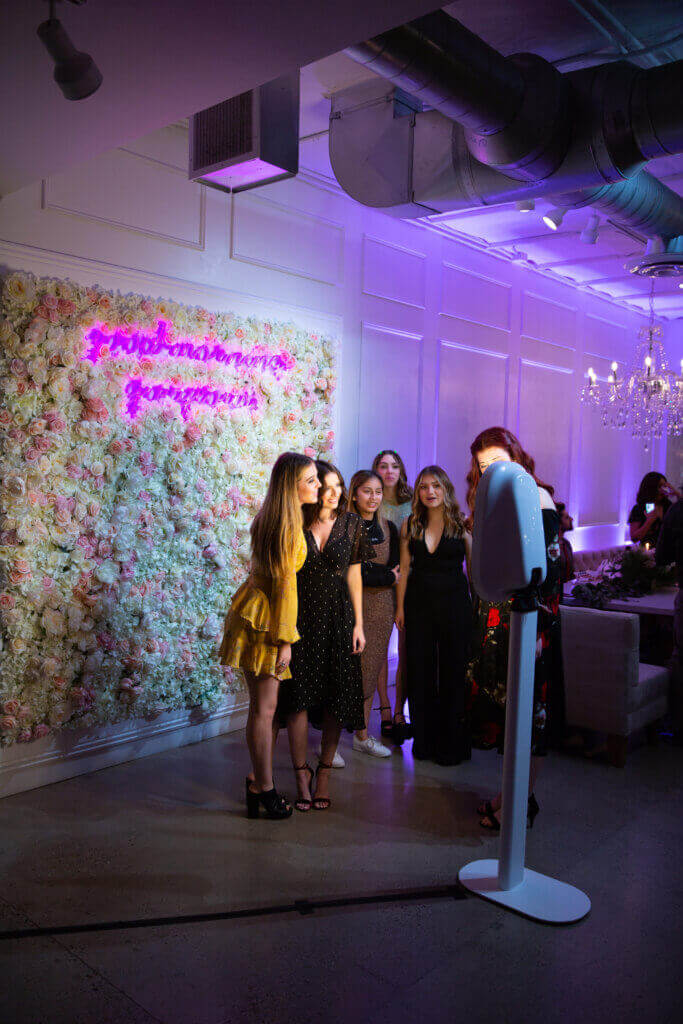 A group of women pose for a photo booth in front of a flower wall with pink neon writing at an indoor event under purple lighting.