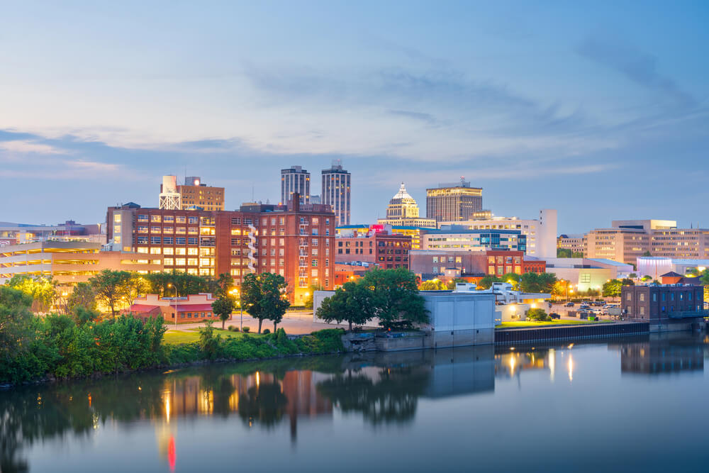 City skyline at dusk with illuminated buildings reflected in a calm river, trees lining the waterfront, and a soft blue sky overhead.
