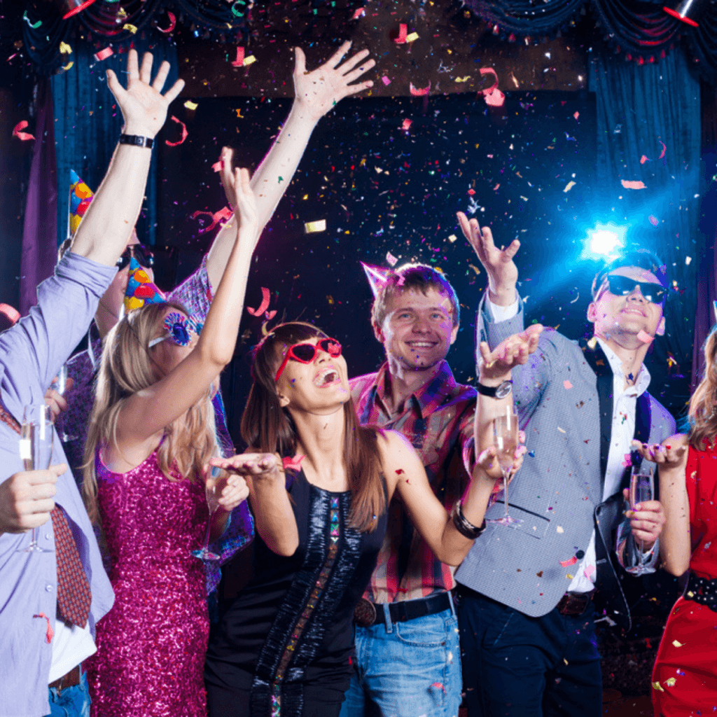 A group of people celebrate at a party, throwing confetti, smiling, and wearing festive clothing and accessories under colorful lights.