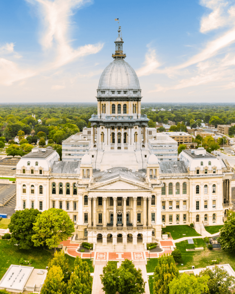 Aerial view of a domed government building with columns, landscaped grounds, and surrounding trees under a partly cloudy sky.